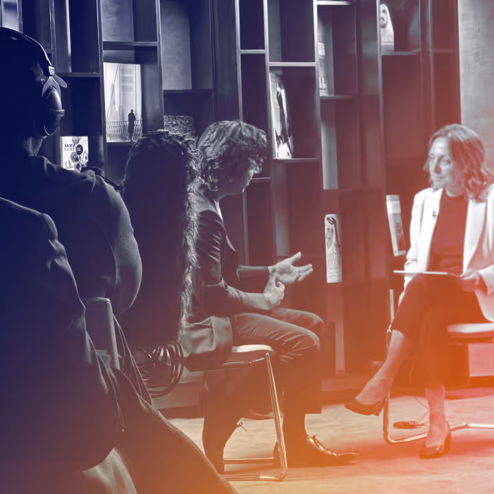 Two people seated and conversing in a modern room with bookshelves, with an audience in the foreground.