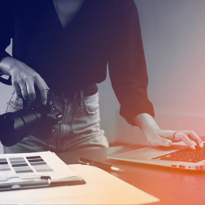Person holding a camera in one hand and using a laptop with the other at a desk with papers and pens.