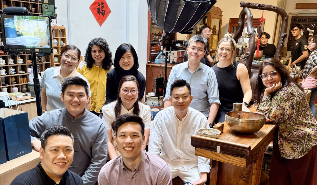 Group of smiling people in casual attire posing indoors around wooden table and shelves.