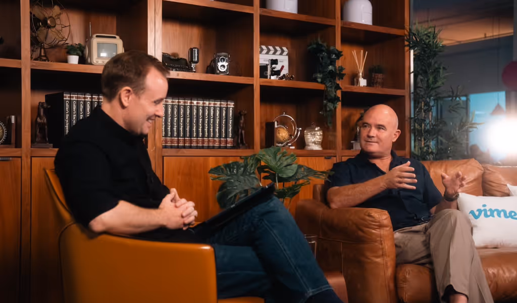 Two men sitting in leather chairs having a conversation in a warmly lit room with wooden shelves.
