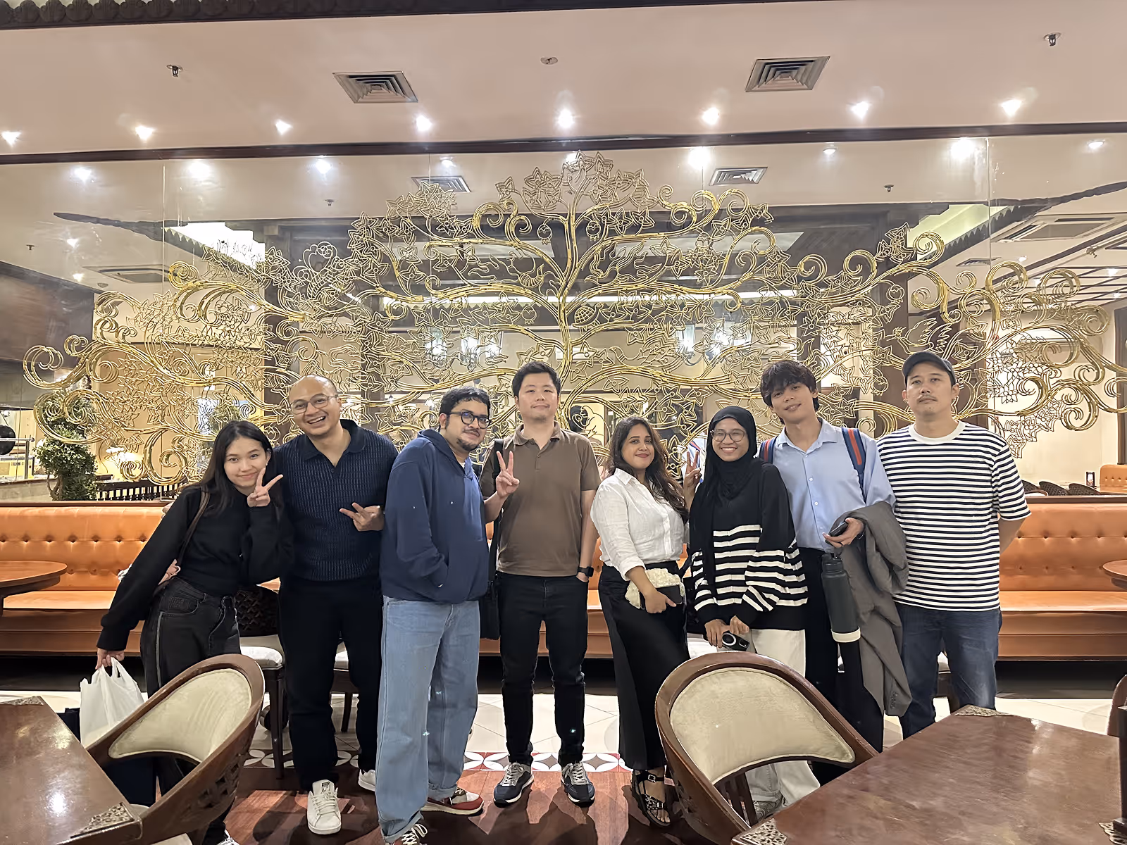 Eight diverse people standing indoors in front of a decorative glass wall with a golden tree design.