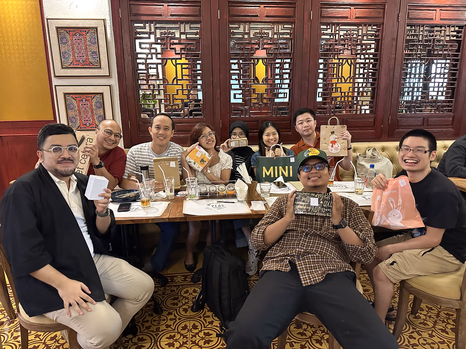 Group of nine people sitting at a table holding gift bags and smiling in a decorated room.