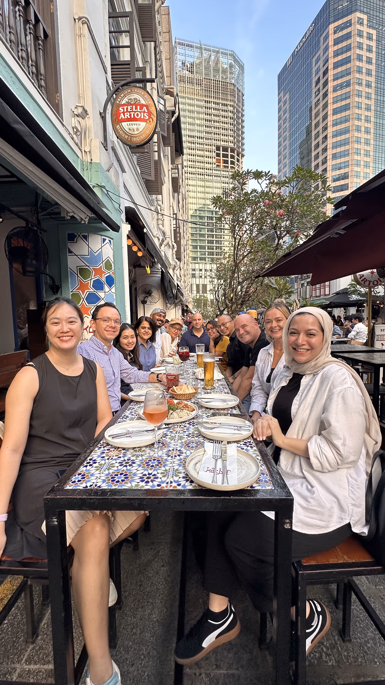 Group of diverse people smiling and seated around a long outdoor mosaic-tiled table in a city setting.