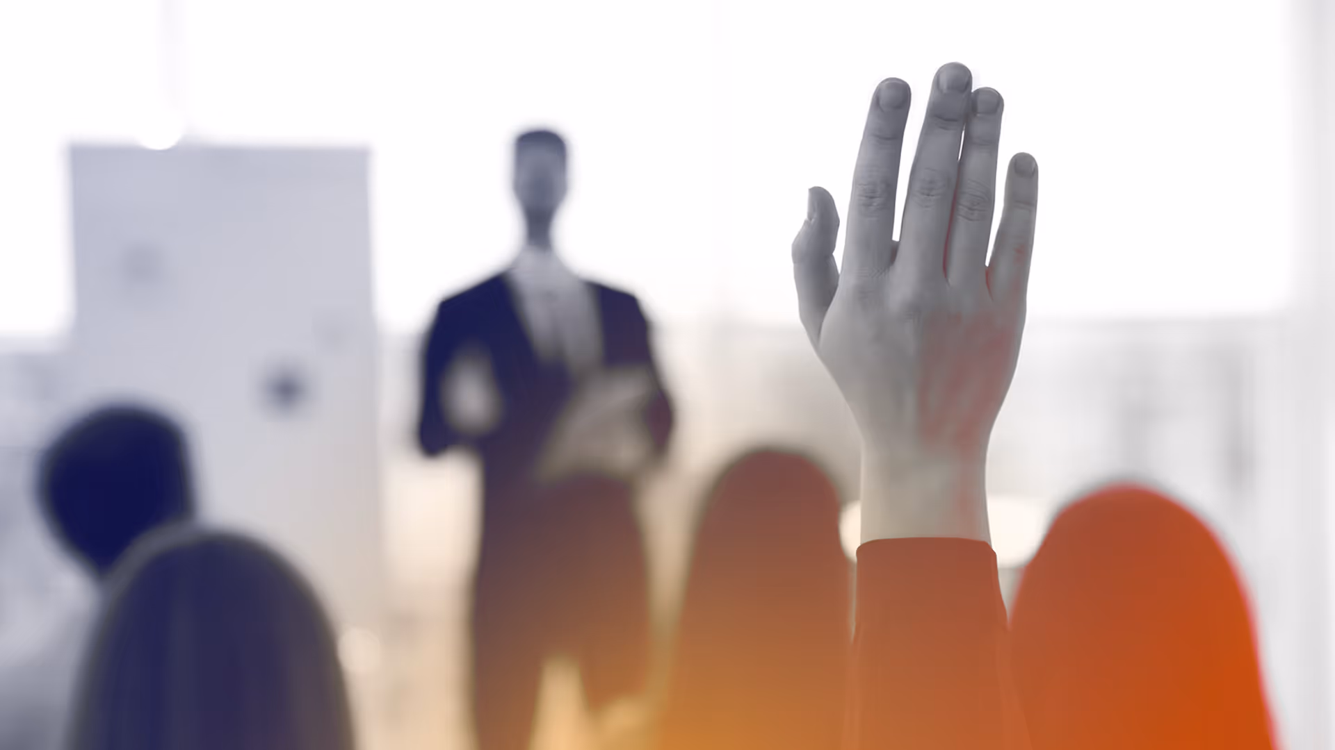Raised hand in a meeting with a speaker in a suit and audience blurred in the background.