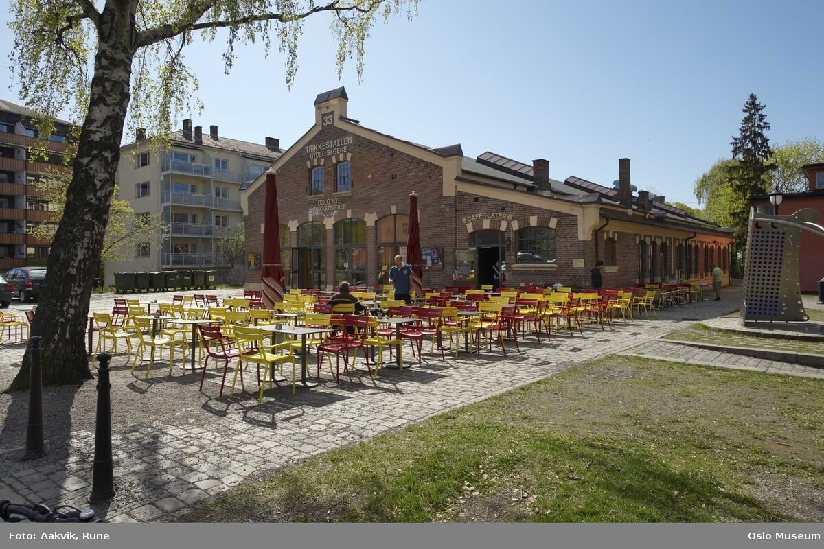 Teaterhuset Trikkestallen på Torshov, et langt murhus med store buede vinduer, fotografert en solskinnsdag med mange bord og stoler på den brosteinbelagte plassen foran inngangen