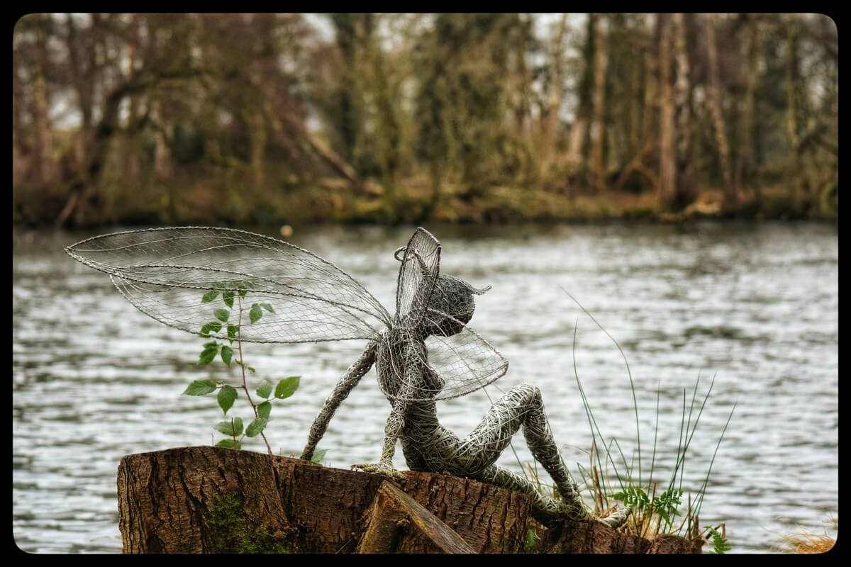 Metallskulptur av alvelignende figur med store vinger laget av netting, sittende på en trestubb ved vannkanten. Naturen i bakgrunnen gir en eventyrlig og magisk stemning