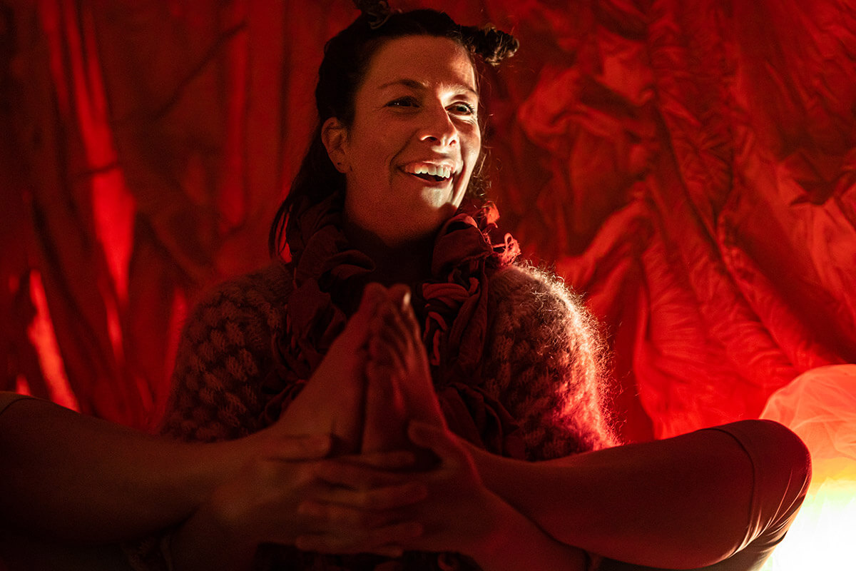 En skuespiller smiler varmt i et mykt, rødt lys under forestillingen «Liv i mor» på Sentralen