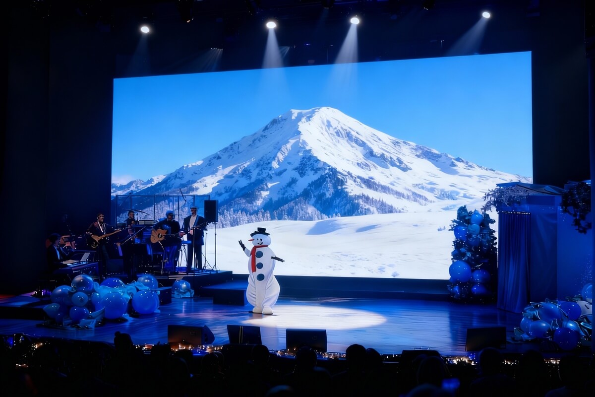 En snømannfigur står på scenen foran et stort bilde av snødekte fjell under en forestilling med musikere i bakgrunnen som illustrasjonsbilde til beskrivelsen av teater og familieforestillinger på Plassen kulturhus i Molde