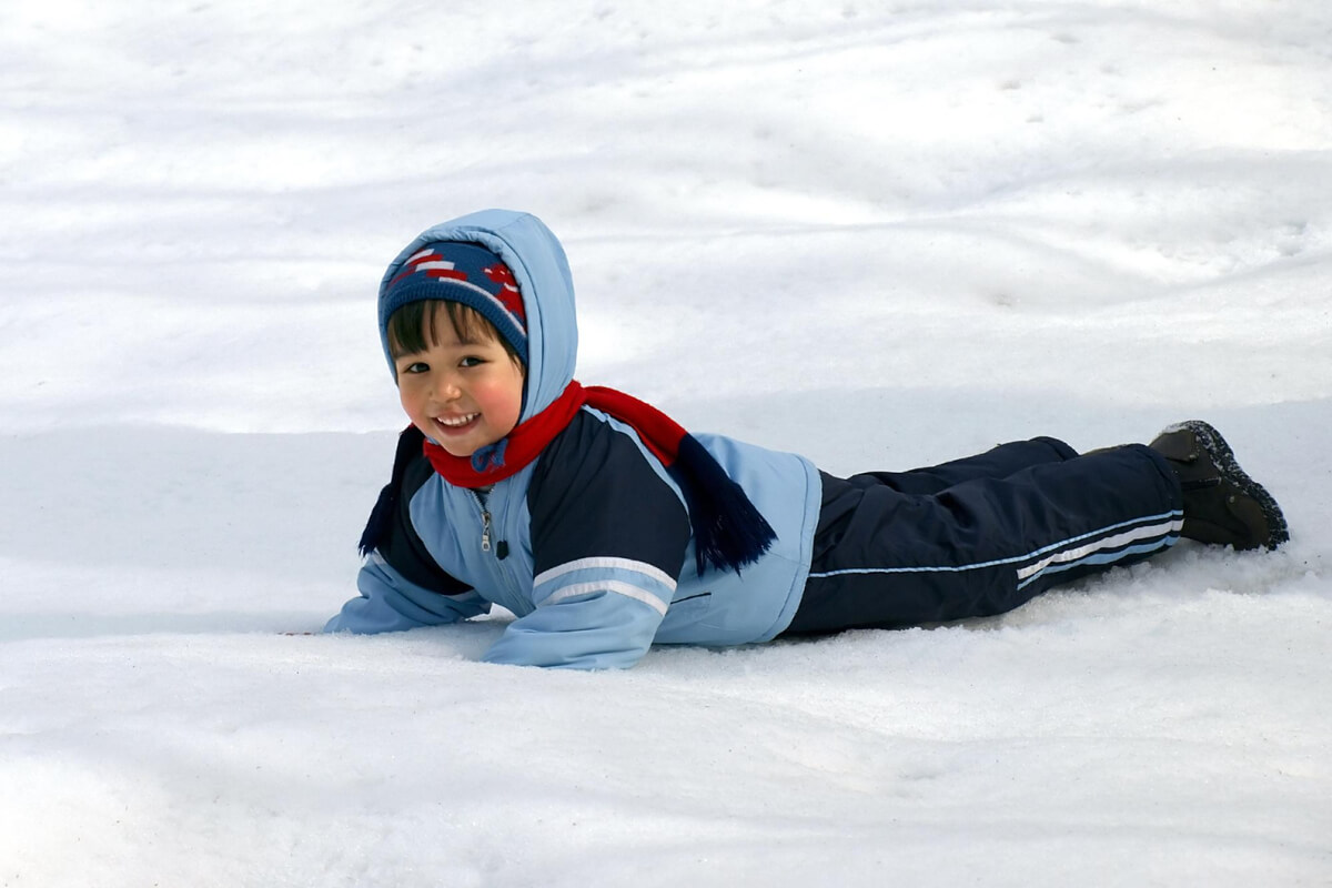 Gutt med blå jakke og svarte bukser som ligger på magen på snøen og smiler som illustrasjon for Kom deg ut-dagen på Lindeberg med DNT