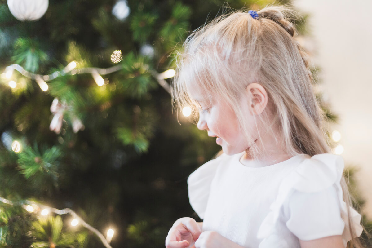 Liten jente i hvit kjole ser ned og smiler foran et pyntet juletre med lys