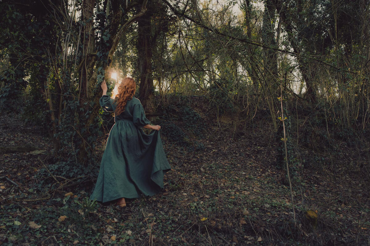 Kvinne i grønn kjole går inn i en mystisk skog med sollys i bakgrunnen – stemningsfull scene fra eventyrteater eller fantasiforestilling i naturen.