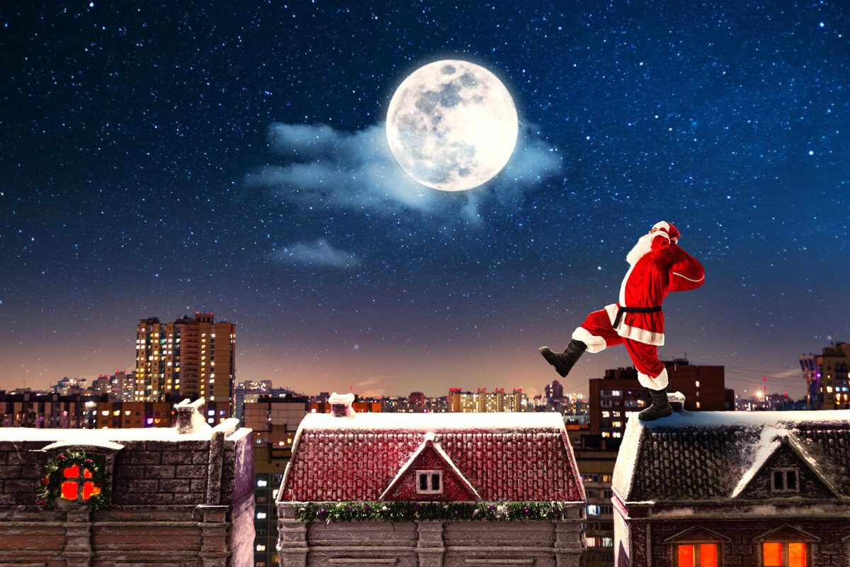 En person i julenissedrakt går over snødekte hustak under en klar nattehimmel med fullmåne