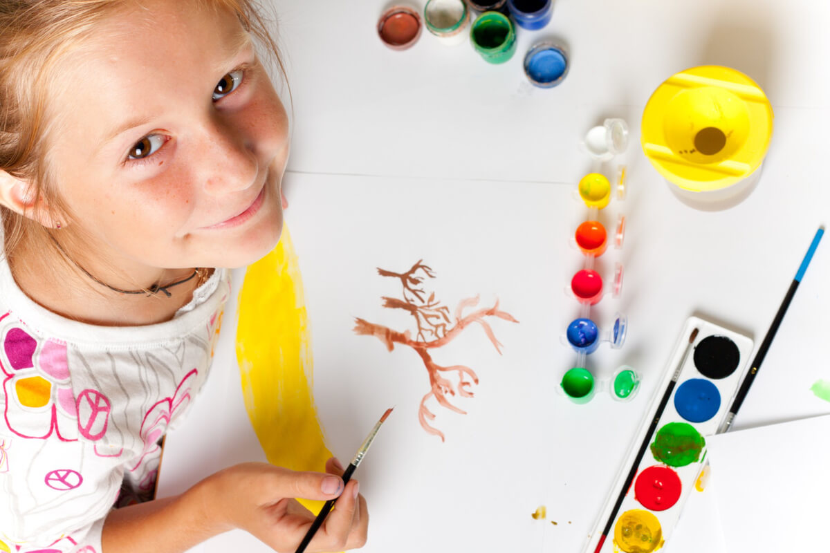 Et barn smiler til kameraet mens hun maler et tre med vannfarger på et ark