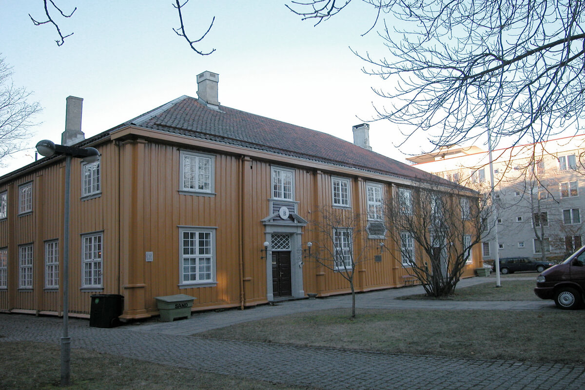 Lagmannsrettsbygningen på Kalvskinnet i Trondheim, et to etasjers gult hus med en leilighetsblokk i bakgrunnen som illustrasjonsbilde for byvandring på Kalvskinnet med NTNU Vitenskapsmuseet