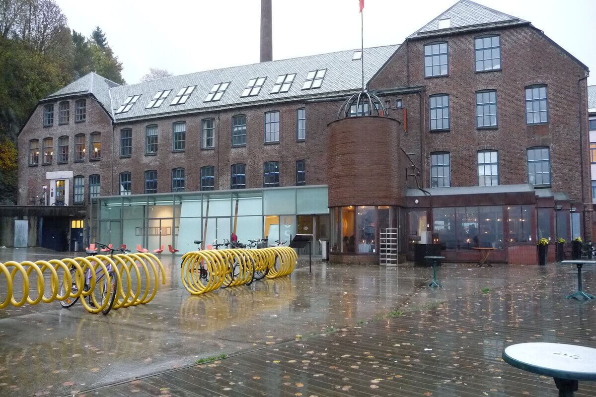 USF Verftet, en bred og høy mursteinsbygning med noen glassfasader på bakkeplan og en stor plass utenfor med gule sykkelstativer som illustrasjon for familiedager på USF Verftet