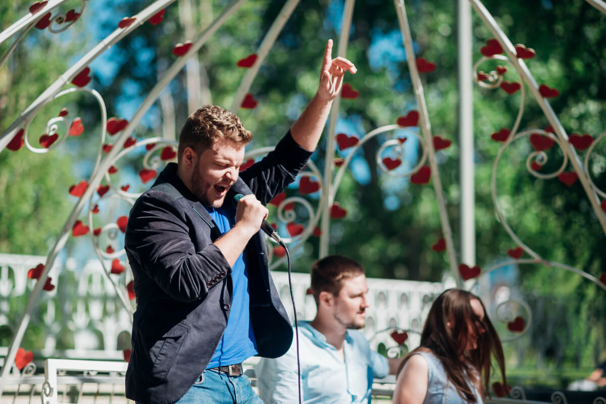 Mann synger med mikrofon og løftet arm under utendørskonsert, med publikum og dekorert paviljong i bakgrunnen