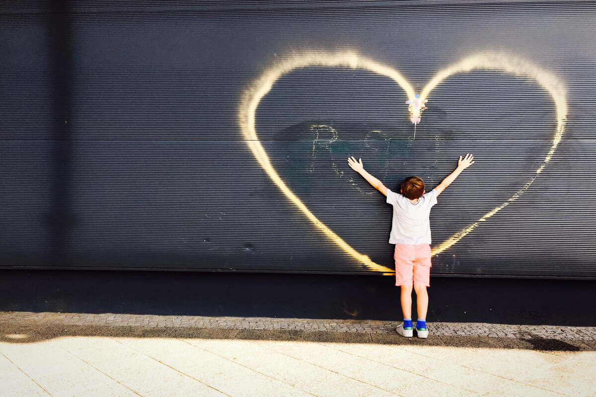 Et barn står foran en mørk vegg med et stort hjerte tegnet i lys, med armene strukket ut mot hjertet som illustrasjonsbilde for beskrivelsen av forestillingen Forelska på Det Andre Teatret