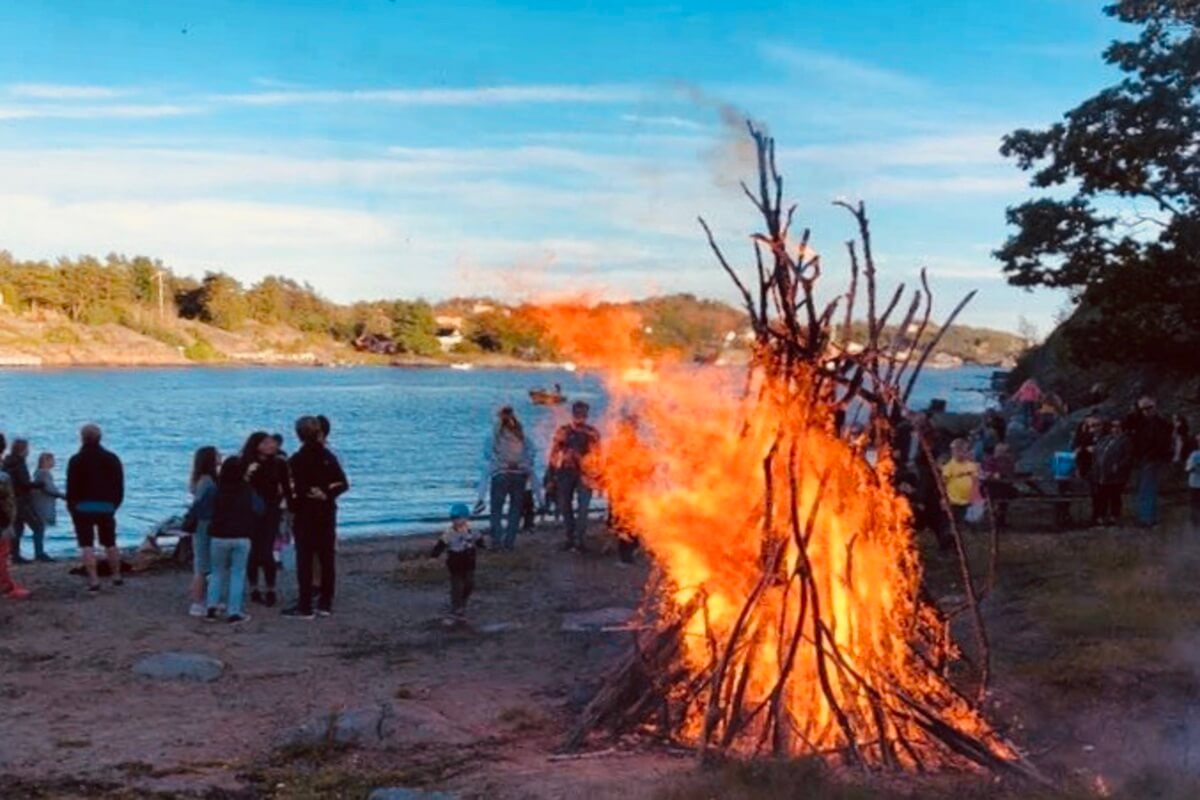 St. Hans-feiring ved sjøen med mennesker samlet rundt et stort bål på stranden, omgitt av vann, skog og kveldshimmel i sommerlys.
