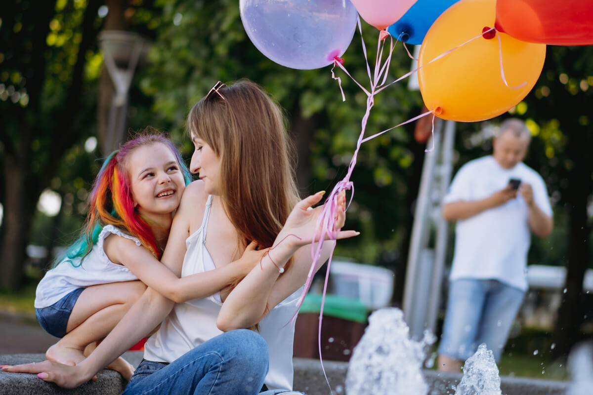 Smilende jente med regnbuefarget hår klemmer moren sin mens de holder ballonger ved en fontene utendørs
