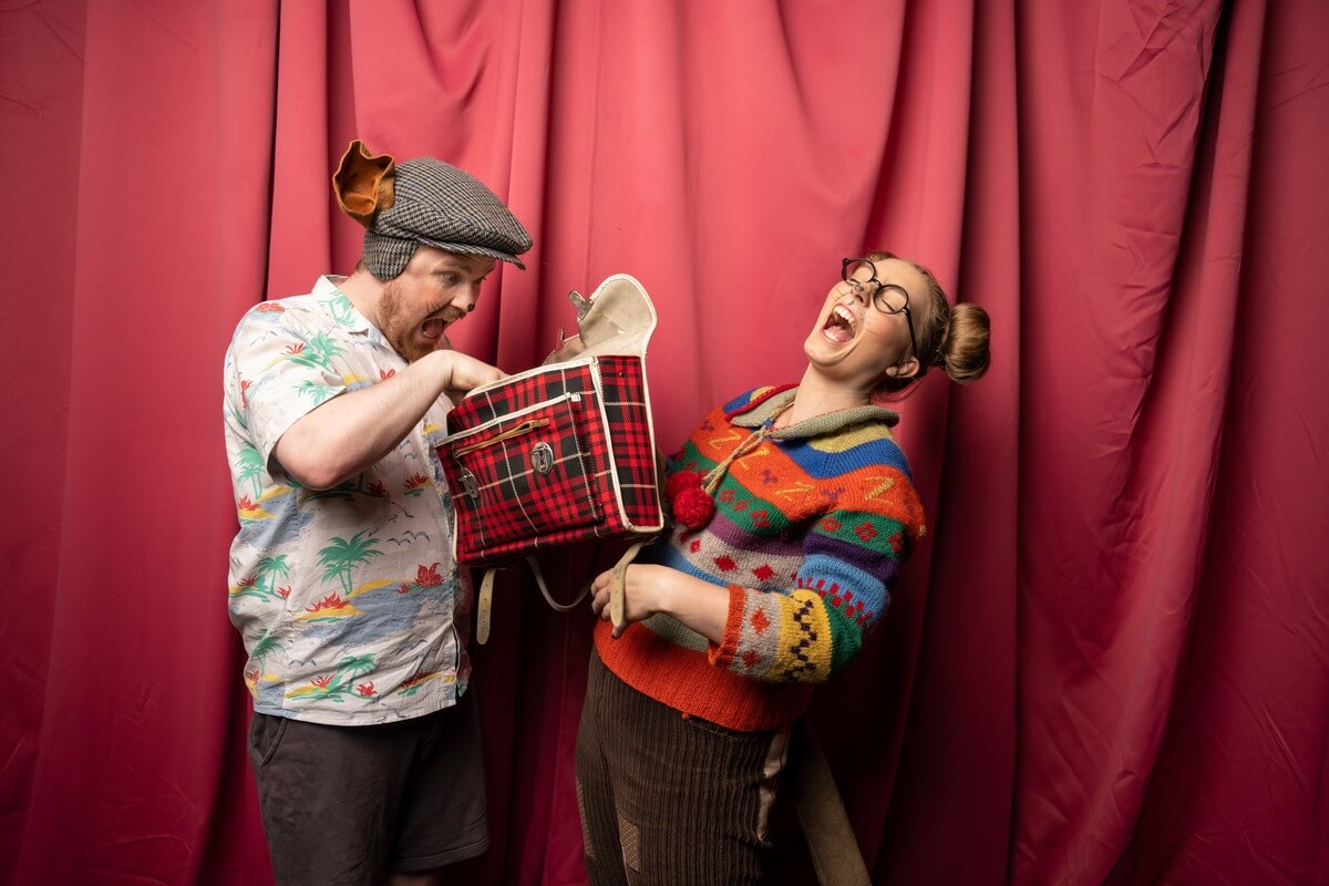 To kostymekledde skuespillere foran rødt sceneteppe, en mann kikker dramatisk ned i en piknikkurv mens en kvinne ler høyt, som illustrasjonsbilde for beskrivelsen av Teaterlørdag på Arbeideren kulturhus