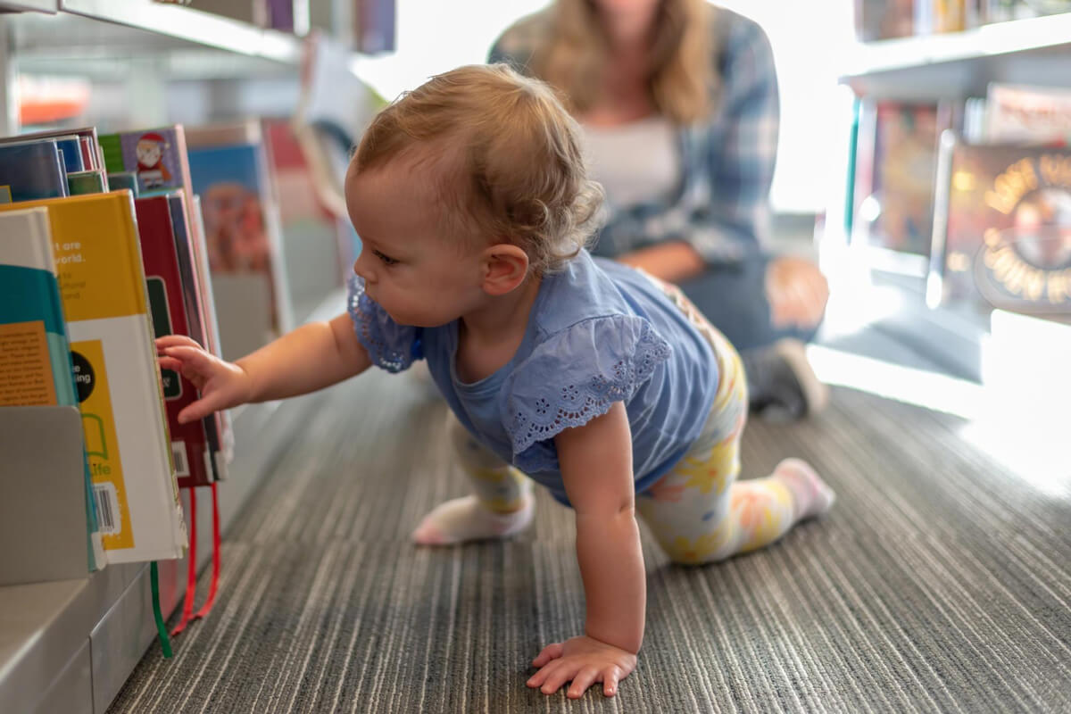 Baby som kravlende utforsker barnebøker på biblioteket mens en voksen følger med i bakgrunnen som illustrasjonsbilde for beskrivelsen av baby på bib på Furuset bibliotek