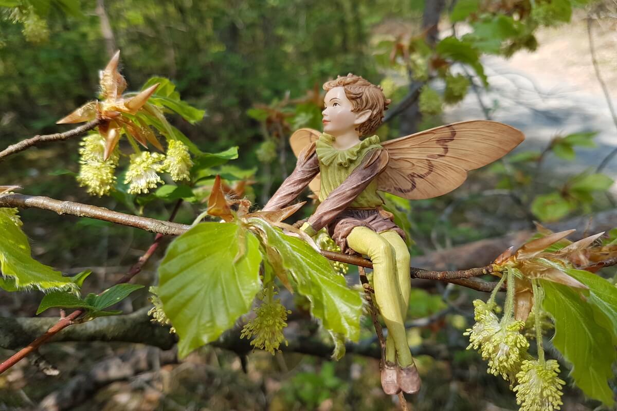 Liten figur av en eventyr- eller alv-lignende skapning med vinger, sittende på en grein med grønne blader og blomsterknopper i en solrik skog