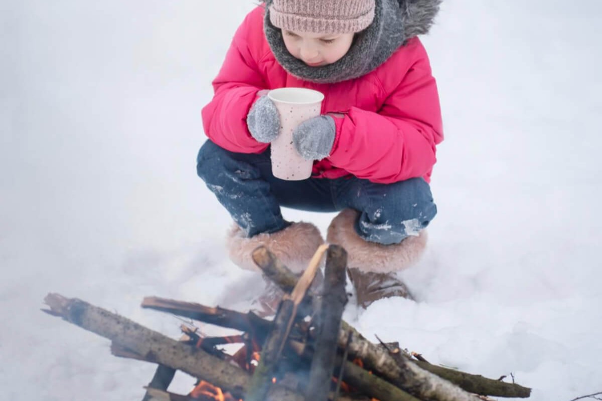 Gutt med rød boblejakke, lue og votter som holder på en kopp foran et bål som illustrasjonsfoto for beskrivelsen av gratis familieskidag med OBOS på Eid Gåd i Asker