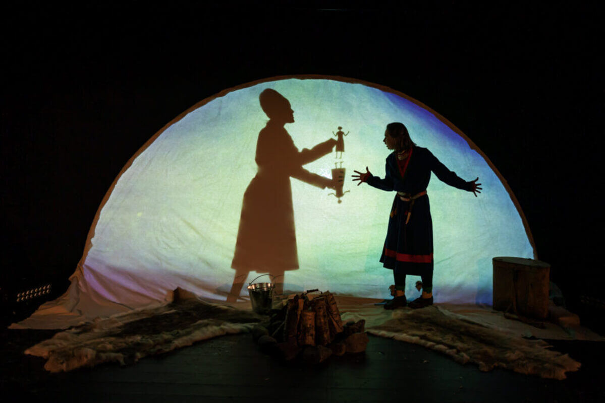 Scene fra teaterforestillingen Dålle, med en skuespiller i samisk drakt foran et opplyst telt som viser skyggeteater med en kvinneskikkelse og figurer.