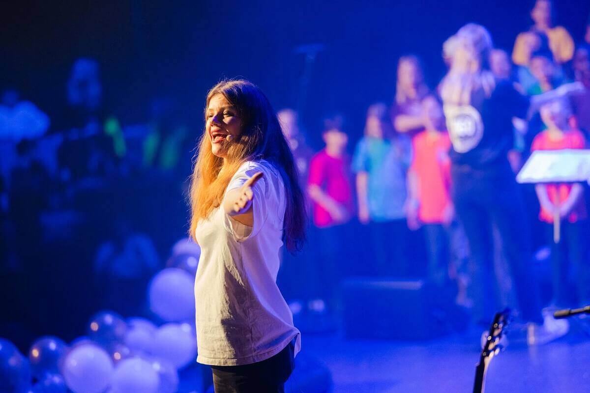 SaraLy syngende på en scene innhyllet i blått lys og med et barnekor i bakgrunnen som illustrasjon til konserten med SaraLy og Unge Oslo på Chateau Neuf