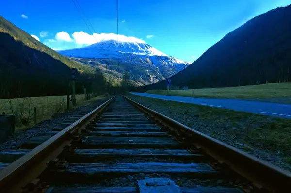 Bilde tatt mellom sporene på en rett strekning på Rjukanbanen med skyggebelagte fjell på siden og Gaustatoppen og blå himmel i bakgrunnen