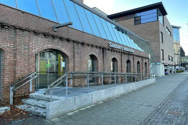 Inngangspartiet til Sandnes bibliotek, rullestolrampe og trapp som går opp til glassdørene i mursteinsbygningen 