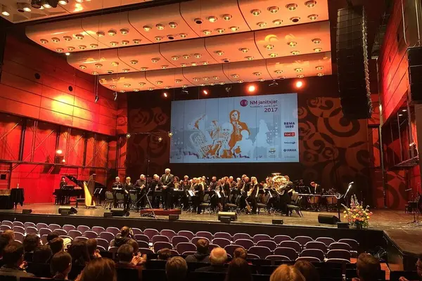 Et janitsjarorkester på scenen under NM Janitsjar 2017, med publikum i salen og stor skjerm i bakgrunnen