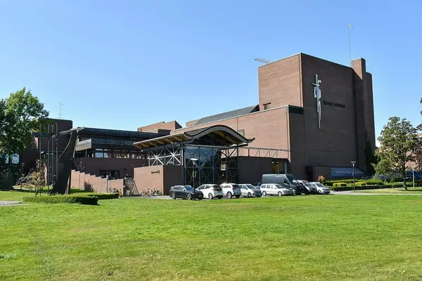 Stor bygningsmasse rød murstein i forskjellige høyder og et buet tak over inngangspartier, og et stort grøntområde foran, som rommer Hjertnes kulturhus  