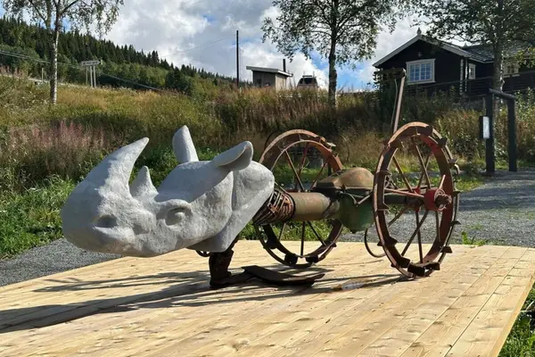 kulptur av et neshorn laget av metall og stein, plassert utendørs på en treplattform ved Eker gård i Valdres