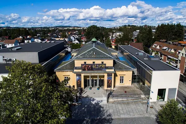 Forsiden med inngangspartiet til Lillestrøm Kultursenter, en eldre bygning med saltak og gulmalt, fotografert fra avstand skrått ovenfra
