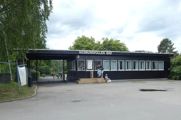 Inngangspartiet til Nebbursvollen friluftsbad med gitterport og en etasjes administrasjonbygg i brunt med mange vinduer og Nebbursvollen bad skrevet med store bokstaver øverst på en del av veggen