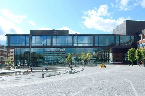 Hamar kulturhus sett fra forsiden, en toetasjes bygning med nesten heldekkende glassfasade, og en stor brosteinsbelagt plass foran