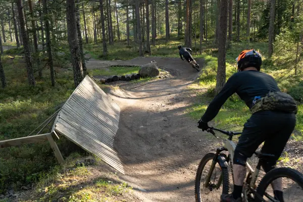 To terrengsyklister nedover Flyten terrengløype i Skimore Drammen BikePark, omgitt av trær og naturlig terreng.