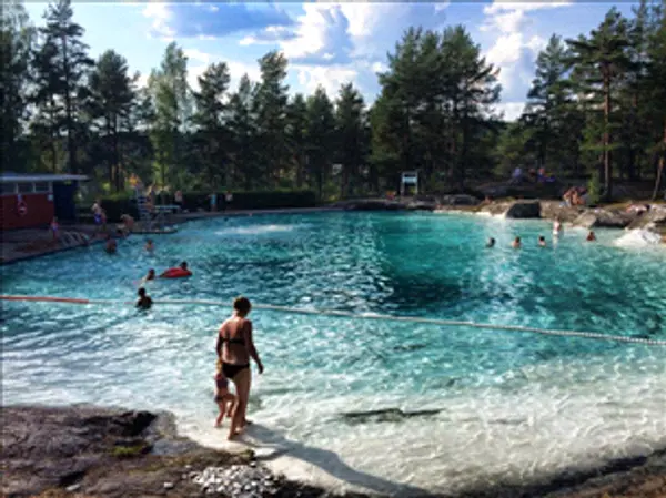 Stort naturlig basseng i fjellet omgitt av spredte trær og med badegjester i vannet på Rånåsfoss Familiebad. 
