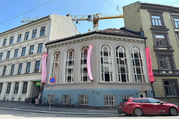 Hjørnehus i betong med blåmalt grunnmur med flere vinduer og toetasjes høye buede vinduer langs begge veggene, som rommer Nordic Black Theatre & Caféteateret