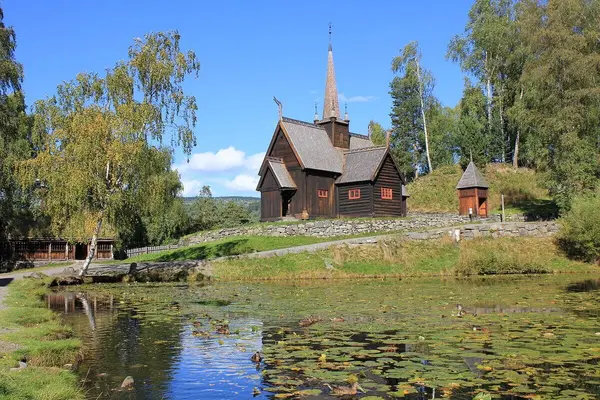 Den gamle kirken på Maihaugen, brunmalt med høyt spir omgitt av trær og med et vann i forgrunnen