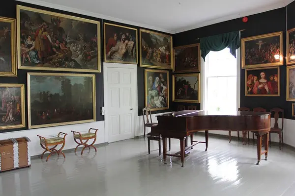  En stue på Gimle Gård med klaver, gråmalt plankegulv og tett i tett med store oljemalerier på veggene