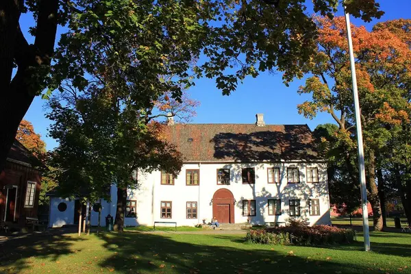 Forsiden på våningshuset på Gjøvik gård, en toetasjes lang hvit murbygning med en stor brun dør midt på med bue over, og stor plen og grønne trær foran