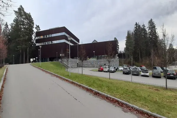 Ankerskogen svømmehall, en stor brun trebygning på en høy steinmur sett mot forsiden, med inngang til venstre og p-plass til høyre