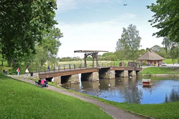 Voldportbroen mellom Gamlebyen i Fredrikstad og landet på andre side av vollgraven, en eldre utseende bro i terreng med grønt gress og trær og et brovokterhus på Gamlebyen siden 