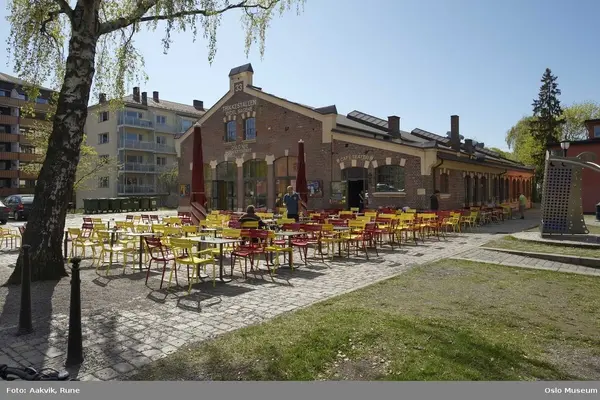Teaterhuset Trikkestallen på Torshov, et langt murhus med store buede vinduer, fotografert en solskinnsdag med mange bord og stoler på den brosteinbelagte plassen foran inngangen