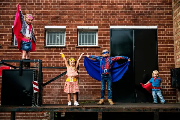 Fire barn i superheltkostymer poserer med armene i været foran en mursteinsvegg som illustrasjonsbilde for beskrivelsen av familiearrangementer og teater for barn i Kulturverket Flammen