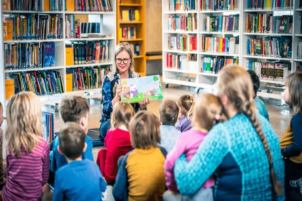 En biblotekar leser høyt fra en billedbok for en gruppe barn i Lørenskog bibliotek, omgitt av fargerike bokhyller. Barna sitter samlet og lytter engasjert mens hun viser boken med illustrasjoner