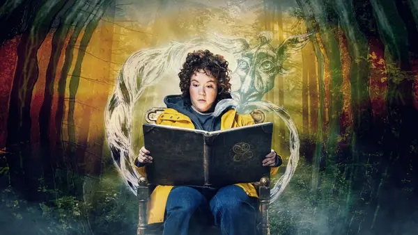 En person i gul jakke sitter og leser i en stor bok, mens en magisk, gjennomsiktig figur av et dyr dukker opp bak dem i en skog som illustrasjonsbilde til barneforestillingen Den uendelige historia på Det Norske Teatret