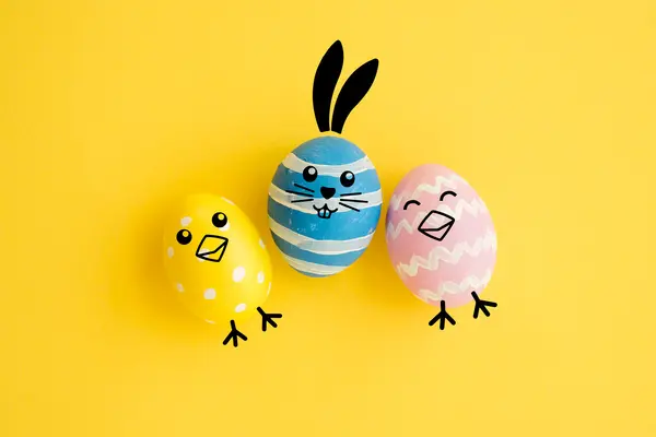 Tre påskeegg dekorert som figurer, med gule og rosa egg som kyllinger og et blått egg som en påskehare mot gul bakgrunn. som illustrasjonbilde for beskrivelsen av påskeaktivitet på Deichman Majorstuen